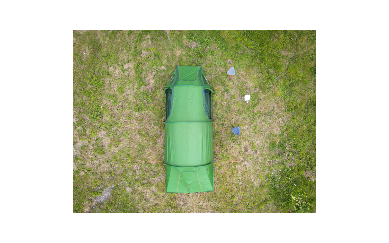 Von oben fotografiertes grünes Tunnelzelt auf einer Wiese; rechts drei kleine dreieckige blaue und graue Gegenstände und ein rundes weißes Objekt
