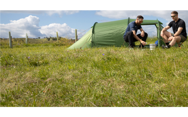 Zwei Personen sitzen vor einem grünen Zelt auf einer Wiese; Holzzaun im Hintergrund; bewölkter Himmel