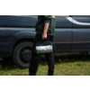 Unterkörper einer Person in grüner Jacke und schwarzer Hose, die eine grün-graue Tasche am Tragegriff hält; dunkler Transporter und Gras im Hintergrund
