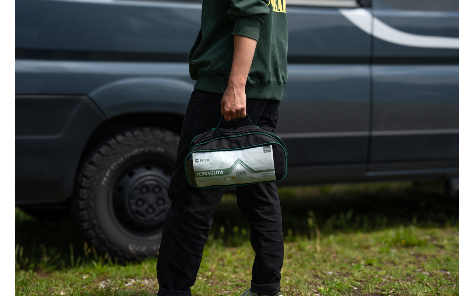 Unterkörper einer Person in grüner Jacke und schwarzer Hose, die eine grün-graue Tasche am Tragegriff hält; dunkler Transporter und Gras im Hintergrund