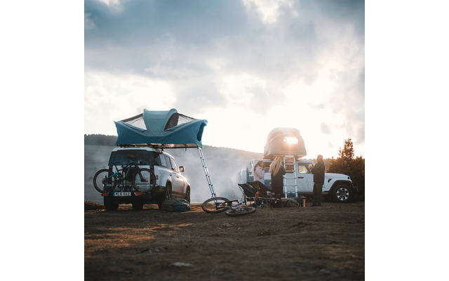Deux VUS avec tentes de toit déployées, vélos fixés à un véhicule, matériel de camping et des personnes sur un terrain découvert, collines et ciel nuageux en arrière-plan