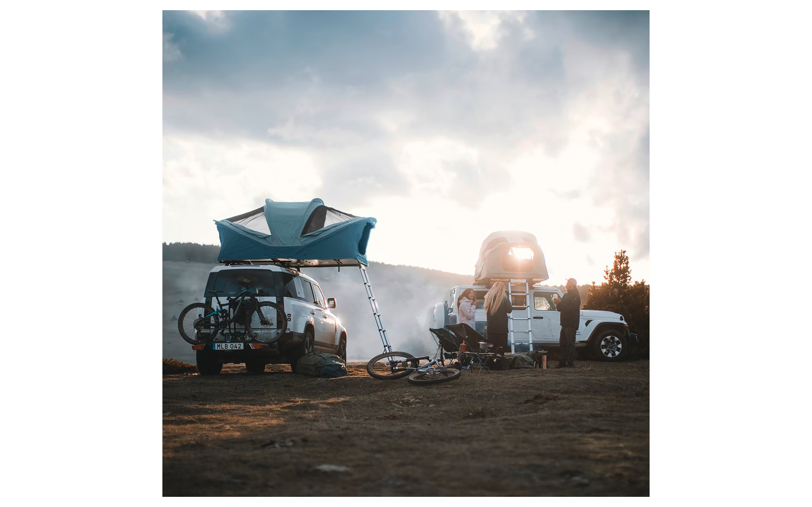 Deux VUS avec tentes de toit déployées, vélos fixés à un véhicule, matériel de camping et des personnes sur un terrain découvert, collines et ciel nuageux en arrière-plan