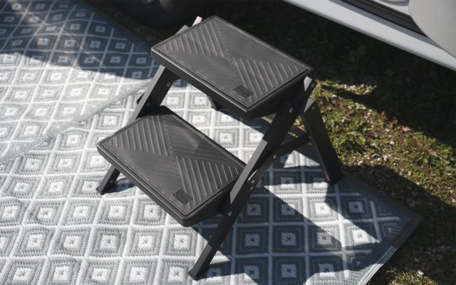 Schwarzer klappbarer Kunststofftritt mit zwei Stufen auf einem grau-weißen, geometrisch gemusterten Teppich, neben Gras und einer offenen Fahrzeugtür, mit Schatten