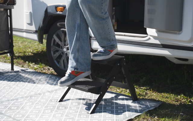 Untere Beine in blauen Jeans und grauen Turnschuhen mit roten Details auf einem schwarzen Klapptritt vor einer offenen weißen Fahrzeugtür, daneben ein Reifen und eine gemusterte Matte auf Gras