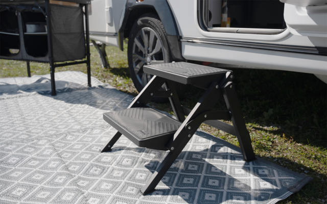 Zweistufige schwarze Klapptrittstufe neben geöffneter Tür eines weißen Campers auf gemustertem Teppich; Gras und Fahrzeugrad sichtbar