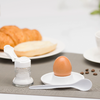 Oeuf brun dans un coquetier blanc sur une petite soucoupe, cuillère blanche à côté, salière ouverte ; en arrière-plan un croissant et un petit pain coupé sur une assiette, tasse de café et grains de café éparpillés sur un set de table gris