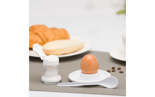 Oeuf brun dans un coquetier blanc sur une petite soucoupe, cuillère blanche à côté, salière ouverte ; en arrière-plan un croissant et un petit pain coupé sur une assiette, tasse de café et grains de café éparpillés sur un set de table gris