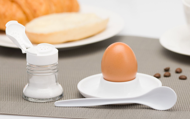 Braunes Ei in weißem Eierbecher auf kleinem Unterteller, weißer Löffel daneben, geöffnete Salzdose; im Hintergrund Croissant und aufgeschnittenes Brötchen auf einem Teller, Kaffeetasse und verstreute Kaffeebohnen auf grauem Tischset