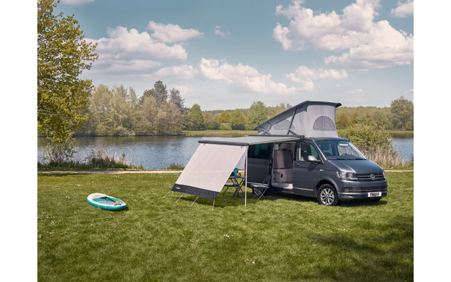 Furgone grigio scuro con tetto a soffietto aperto e tendalino esteso su una riva erbosa; tavola da paddle sull'erba, alberi e cielo nuvoloso