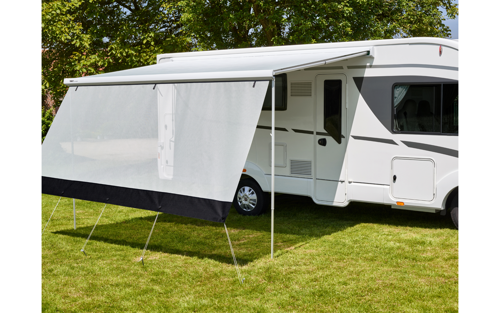 Camper bianco con tendalino aperto e schermo laterale su prato, alberi sullo sfondo