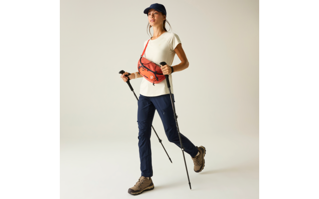 Persoon met donkerblau petje, wit T‑shirt, donkerblau wandelbroek, bruine wandelschoenen, oranje heuptasje schuin over de borst, houdt twee wandelstokken, lichte effen achtergrond