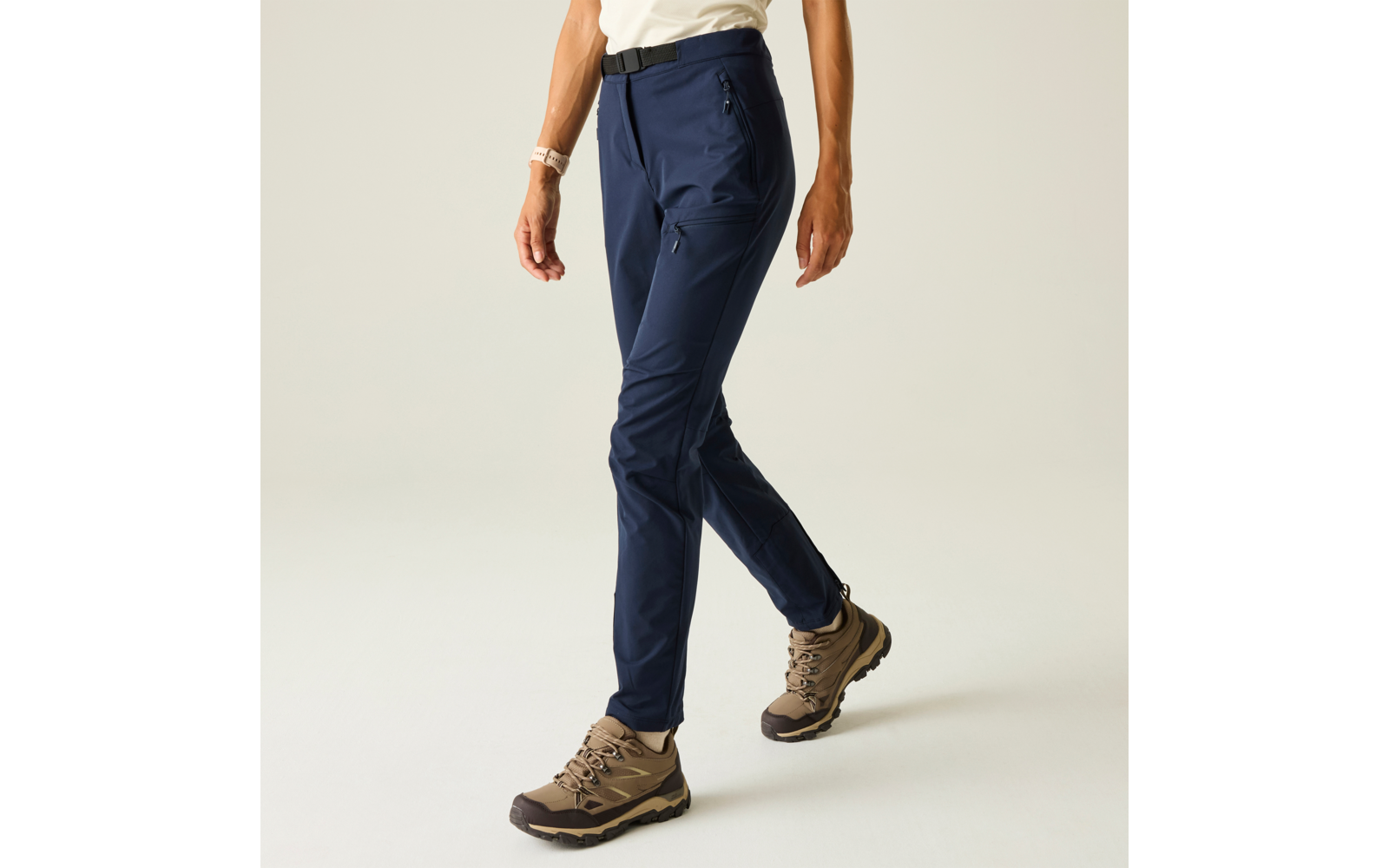 Onderlichaam van een persoon, alleen benen en heupen zichtbaar; donkerblauwe broek, beige-bruin veterschoenen met profielzool; lichte achtergrond