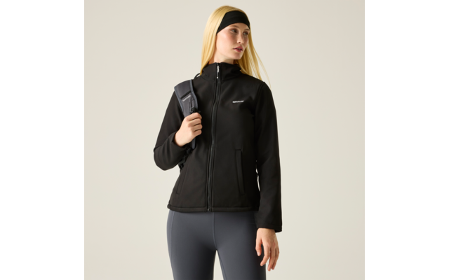 Frau mit langem blondem Haar und Stirnband, trägt schwarze Jacke mit Reißverschluss, graue Leggings und einen Rucksackriemen über einer Schulter vor hellem Hintergrund