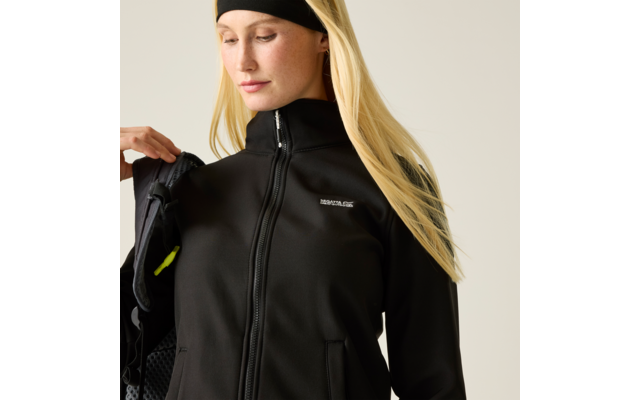 Person mit langen blonden Haaren, schwarzem Haarband und schwarzer Jacke mit Reißverschluss, hält eine Rucksackriemen über einer Schulter und blickt nach unten