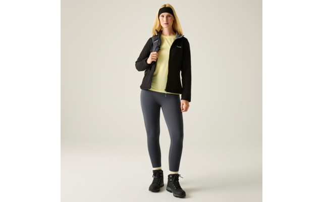 Person frontal stehend, schwarzes Stirnband, schwarze Fleecejacke, hellgelbes T‑Shirt, dunkelgraue Leggings, schwarze knöchelhohe Stiefel, Rucksack über einer Schulter, heller einfarbiger Hintergrund