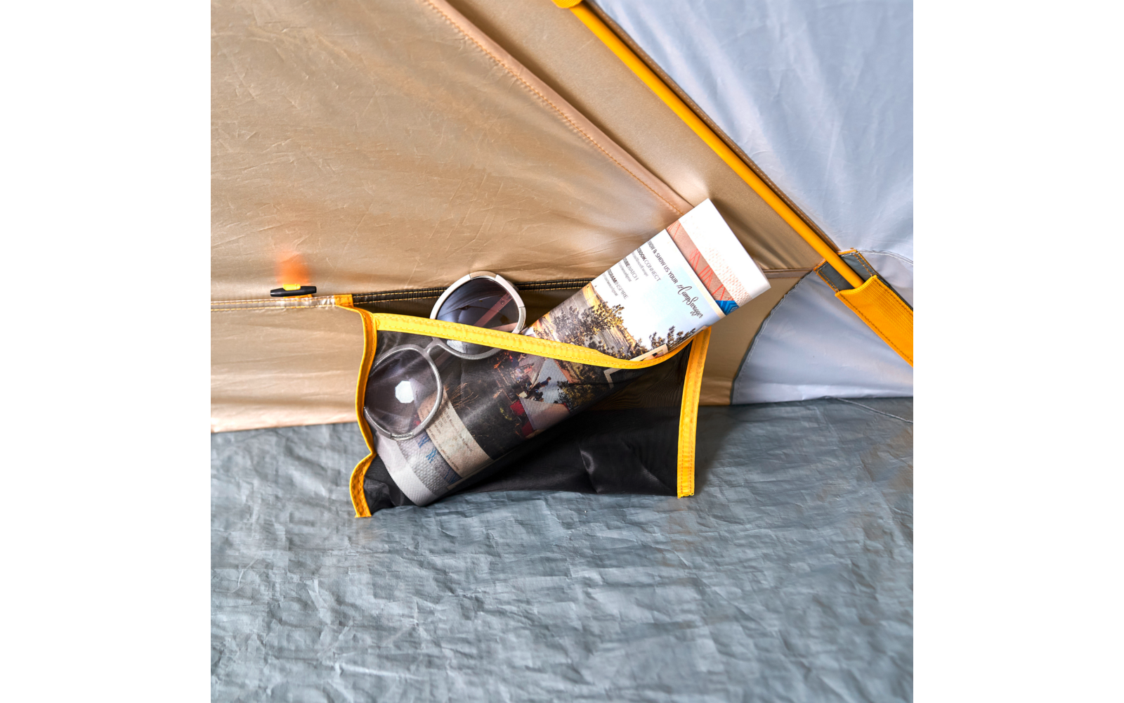 Innenraum eines Zelts mit beiger Innenwand, gelbem Rand und Stofftasche, darin eine aufgerollte Zeitschrift und eine Broschüre auf grauem Boden
