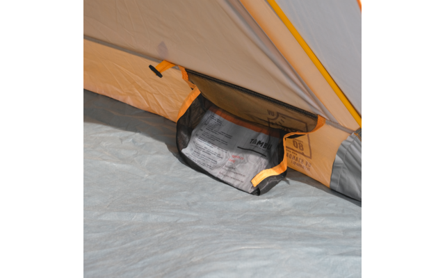 Innenraum eines Zeltes: grauer Boden, beige Innenwand mit orangefarbener Naht und eine schwarze Netzinnentasche, die ein gefaltetes, bedrucktes Papier enthält