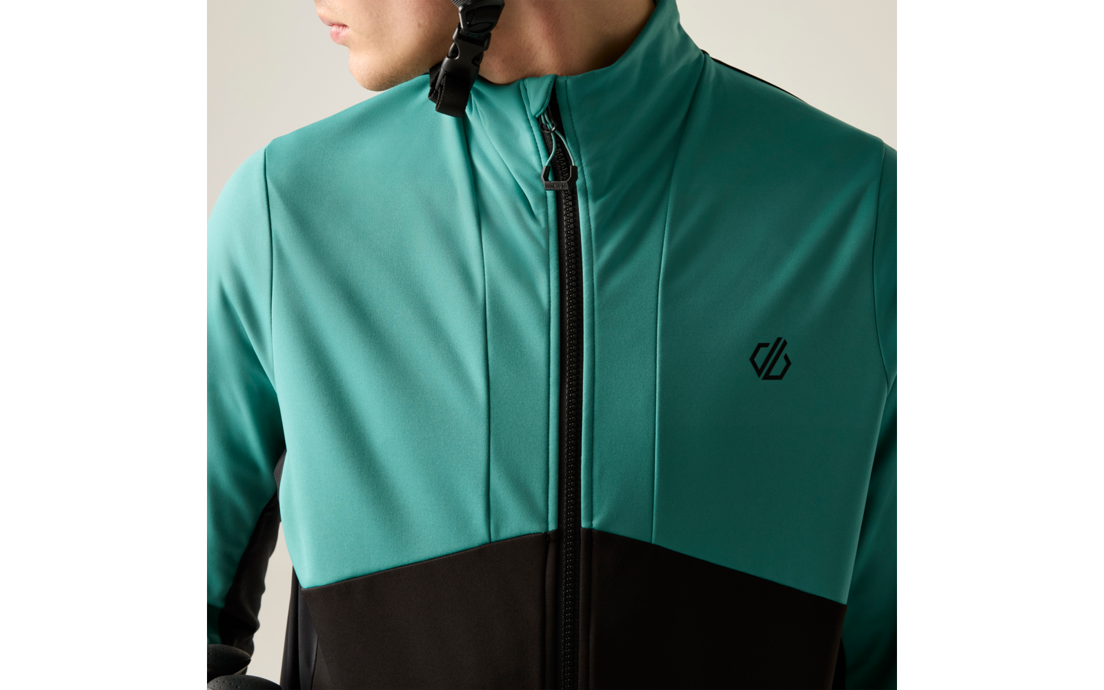 Oberkörper einer Person in türkiser und schwarzer Jacke mit zentralem Reißverschluss und kleinem Logo auf der Brust; Gesicht nur teilweise sichtbar