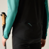 Rückenansicht einer Person von der Taille abwärts, schwarzes Vest mit türkisfarbenen Ärmeln, hält zwei Wanderstöcke mit Korkgriffen in der linken Hand