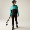 Person in türkis-schwarzer Jacke, schwarzer Hose, Skihelm und Skischuhen, hält gefalteten schwarzen Regenschirm mit Holzgriff, steht vor neutralem Hintergrund und blickt nach links