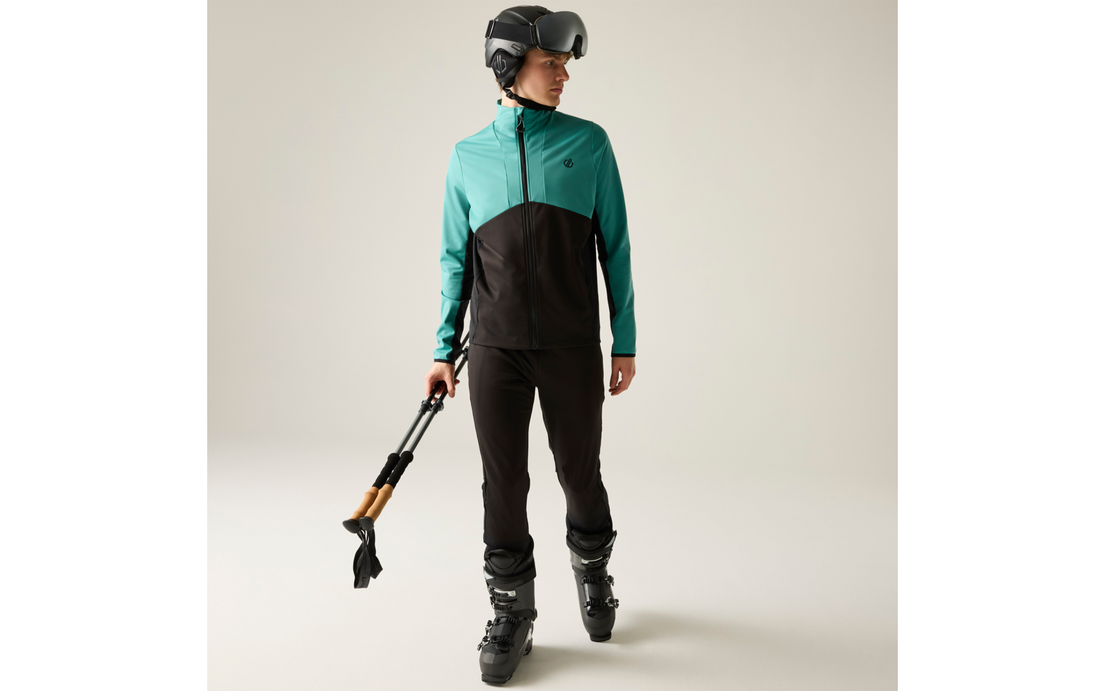 Person in türkis-schwarzer Jacke, schwarzer Hose, Skihelm und Skischuhen, hält gefalteten schwarzen Regenschirm mit Holzgriff, steht vor neutralem Hintergrund und blickt nach links