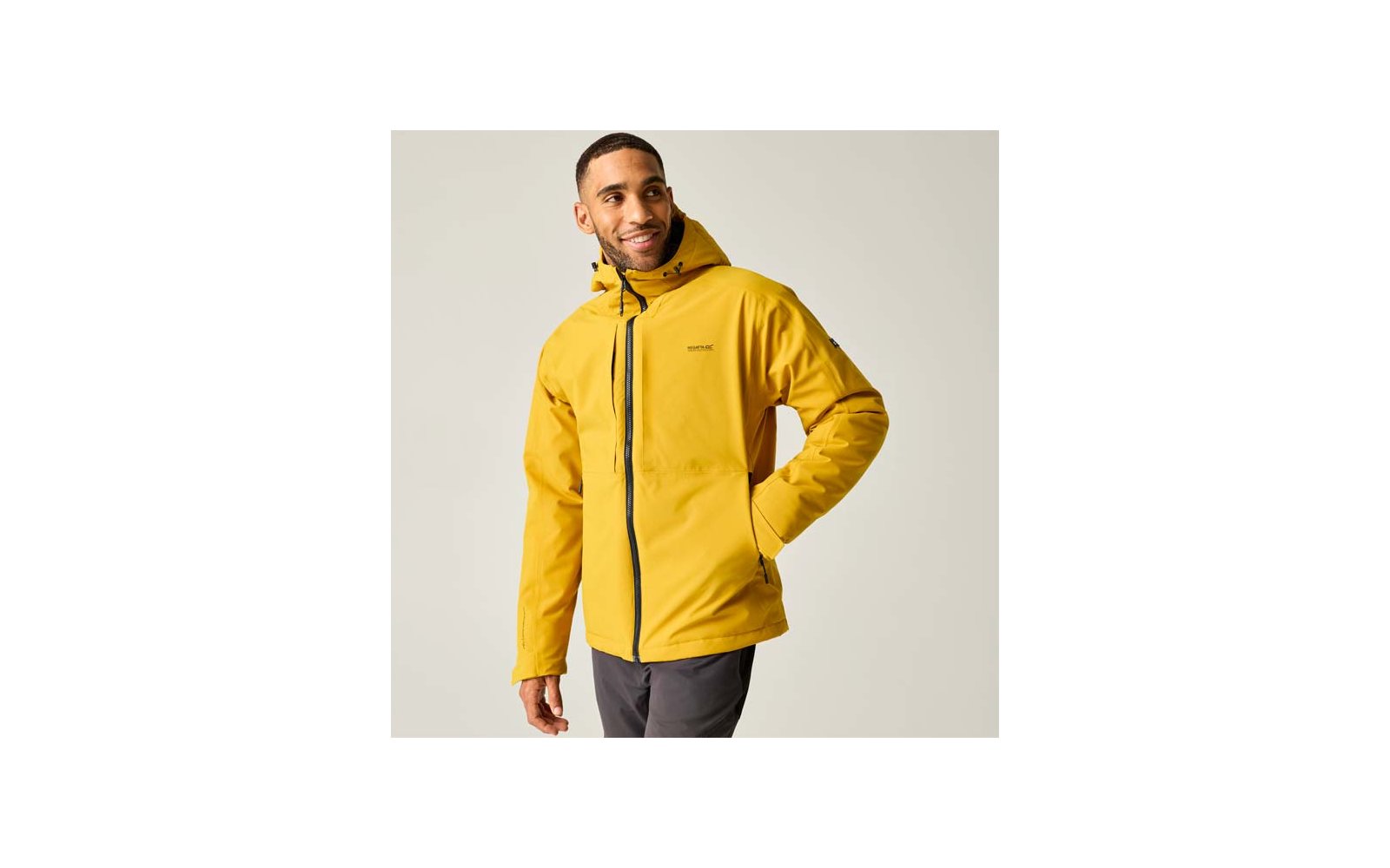Person in gelber Kapuzenjacke mit durchgehendem Reißverschluss, Hand in Seitentasche, dunkle Hose, heller Hintergrund