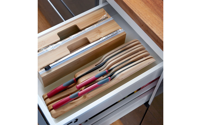 Tiroir blanc ouvert avec insert en bambou ; couteaux empilés aux manches en bois avec zones de préhension rouge-gris ; à gauche deux rouleaux de film aluminium emballés dans du plastique ; bord d’un plan de travail en bois visible