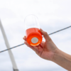 Hand hält ein Glas mit oranger Flüssigkeit vor hellem Himmel