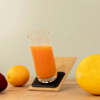 Hohes Glas mit orangefarbener Flüssigkeit auf schräg gestelltem Holzbrett mit schwarzem Untersetzer; links eine Orange und eine dunkelrote Frucht, rechts eine gelbe Melone auf einer Holzoberfläche