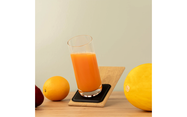 Hohes Glas mit orangefarbener Flüssigkeit auf schräg gestelltem Holzbrett mit schwarzem Untersetzer; links eine Orange und eine dunkelrote Frucht, rechts eine gelbe Melone auf einer Holzoberfläche