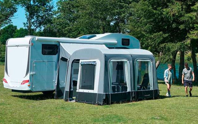 Weißer Wohnwagen mit grauem Vorzelt auf einer Grasfläche neben Bäumen und einem See, zwei Personen gehen am Ufer, blauer Himmel