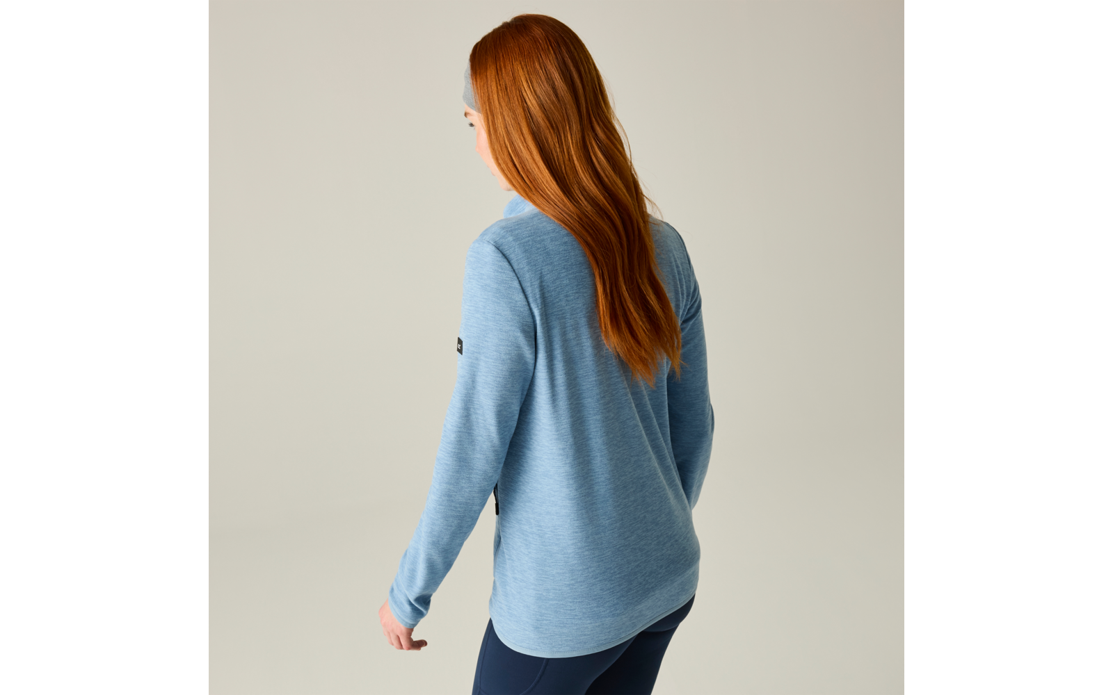 Persona vista da dietro con lunghi capelli rossi, maglia azzurro chiaro a maniche lunghe e pantaloni scuri su sfondo neutro