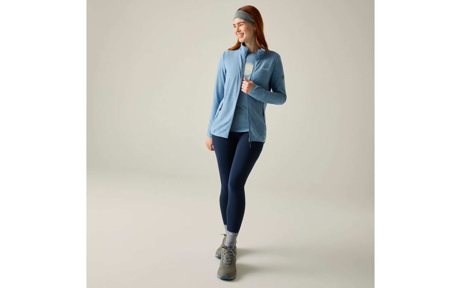 Mujer con chaqueta azul claro, camiseta azul, mallas oscuras, cinta en la cabeza, calcetines grises y botas de senderismo grises sobre fondo claro