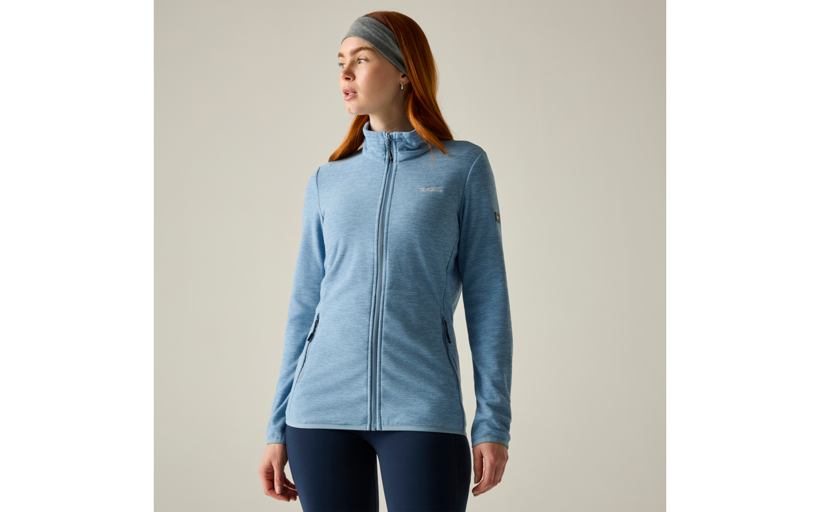 Persona con capelli rossi, fascia grigia, giacca azzurra con cerniera e leggings scuri, in piedi e rivolta verso sinistra su sfondo chiaro