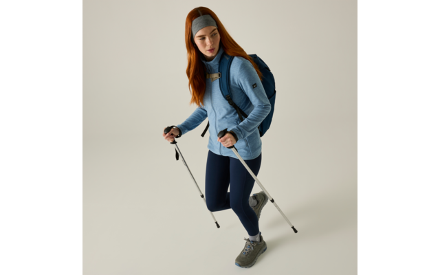 Persona con capelli rossi, fascia grigia, maglione blu, leggings scuri, scarpe da trekking, zaino blu e due bastoncini da trekking su sfondo chiaro