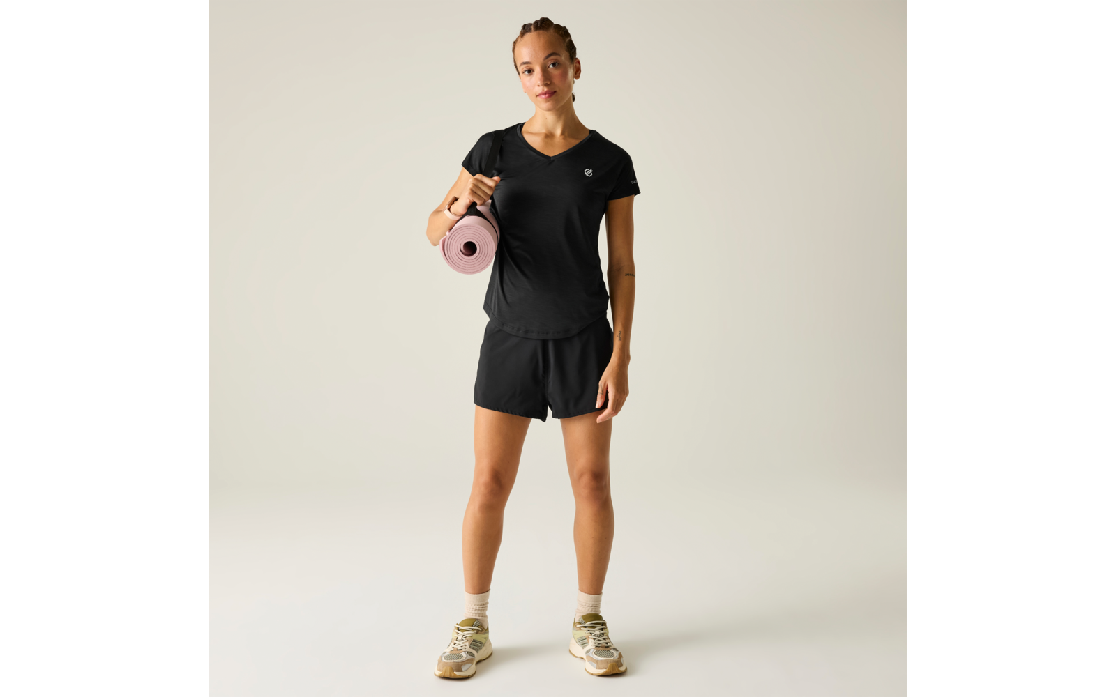 Person steht frontal auf hellem Hintergrund, trägt schwarzes T‑Shirt und schwarze Shorts, hält eine rosa Yogamatte auf der linken Schulter, trägt helle Sneaker und Socken, Haare zu Zöpfen zurückgebunden