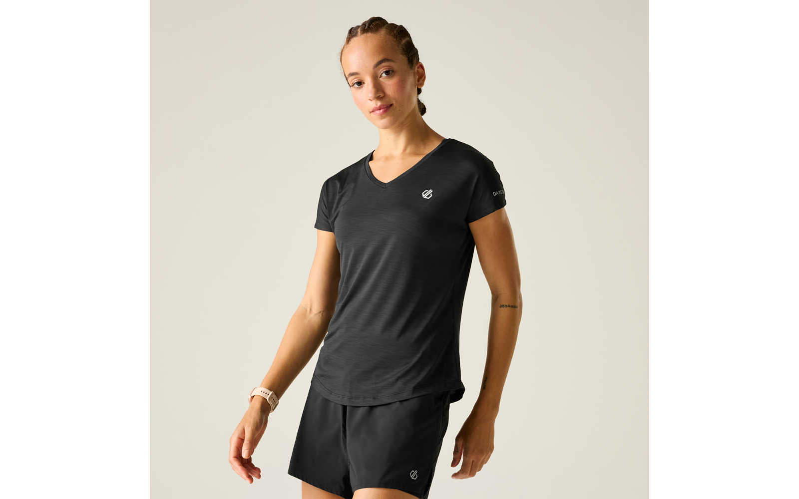 Person mit geflochtenen Haaren, trägt schwarzes V‑Ausschnitt-Sportshirt und schwarze Sportshorts mit weißen Logos, helles Armband am linken Handgelenk, feine Tätowierung am rechten Unterarm, vor hellem einfarbigem Hintergrund