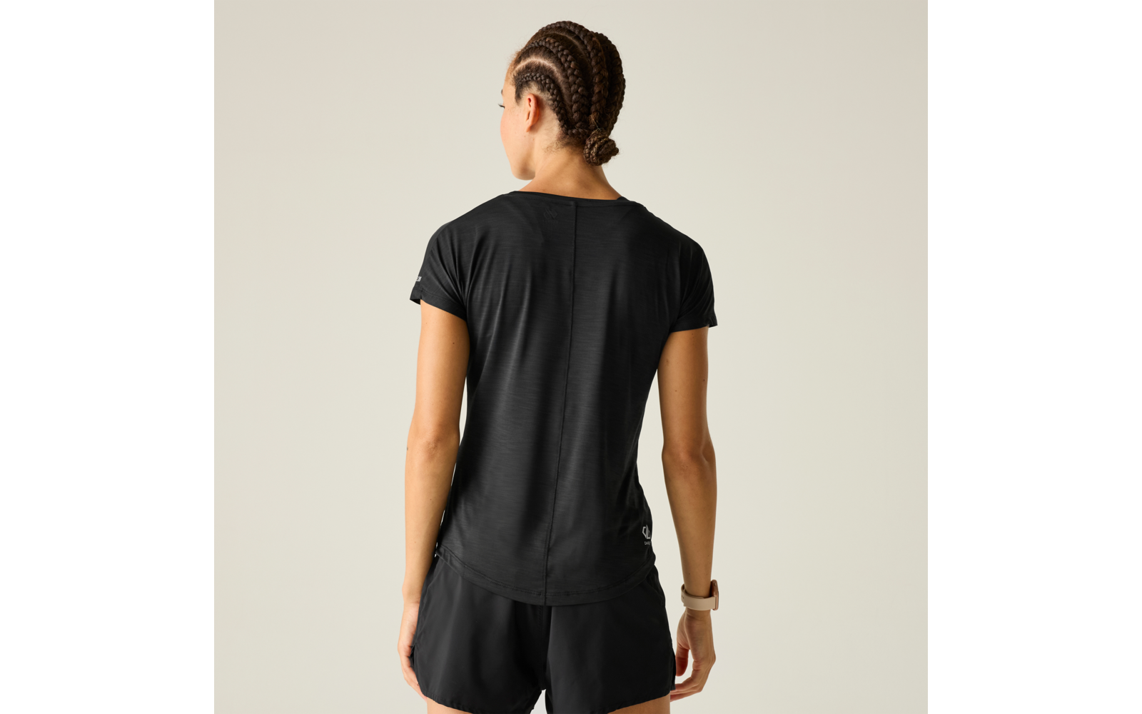 Person von hinten, trägt schwarzes kurzärmeliges T-Shirt und schwarze Shorts, geflochtene Haare zu einem Dutt gebunden, Smartwatch am linken Handgelenk, heller Hintergrund