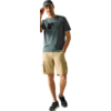 Stehende Person mit Baseballcap, eine Hand an der Krempe, andere Hand in Hosentasche; blaues T‑Shirt mit quadratischem Aufdruck, beige Cargoshorts, schwarze Sneaker