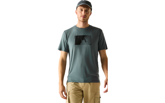 Person steht frontal, trägt graue Kappe, blaugrünes T-Shirt mit schwarzem rechteckigem Motiv auf der Brust, beige Cargoshorts, Hände seitlich, hellbeiger Hintergrund