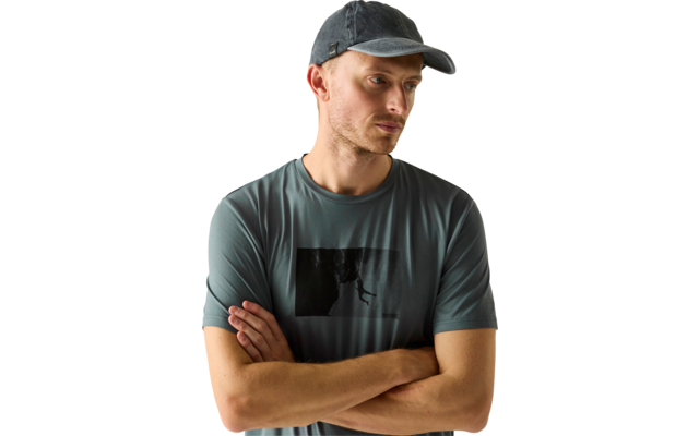 Mann mit Baseballcap und T‑Shirt, Arme verschränkt, Blick nach unten rechts, heller neutraler Hintergrund, dunkles rechteckiges Motiv auf dem T‑Shirt