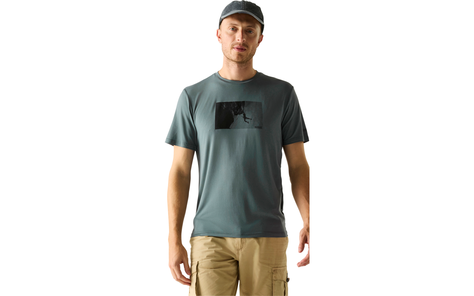 Person steht frontal, trägt graue Kappe, blaugrünes T-Shirt mit schwarzem rechteckigem Motiv auf der Brust, beige Cargoshorts, Hände seitlich, hellbeiger Hintergrund