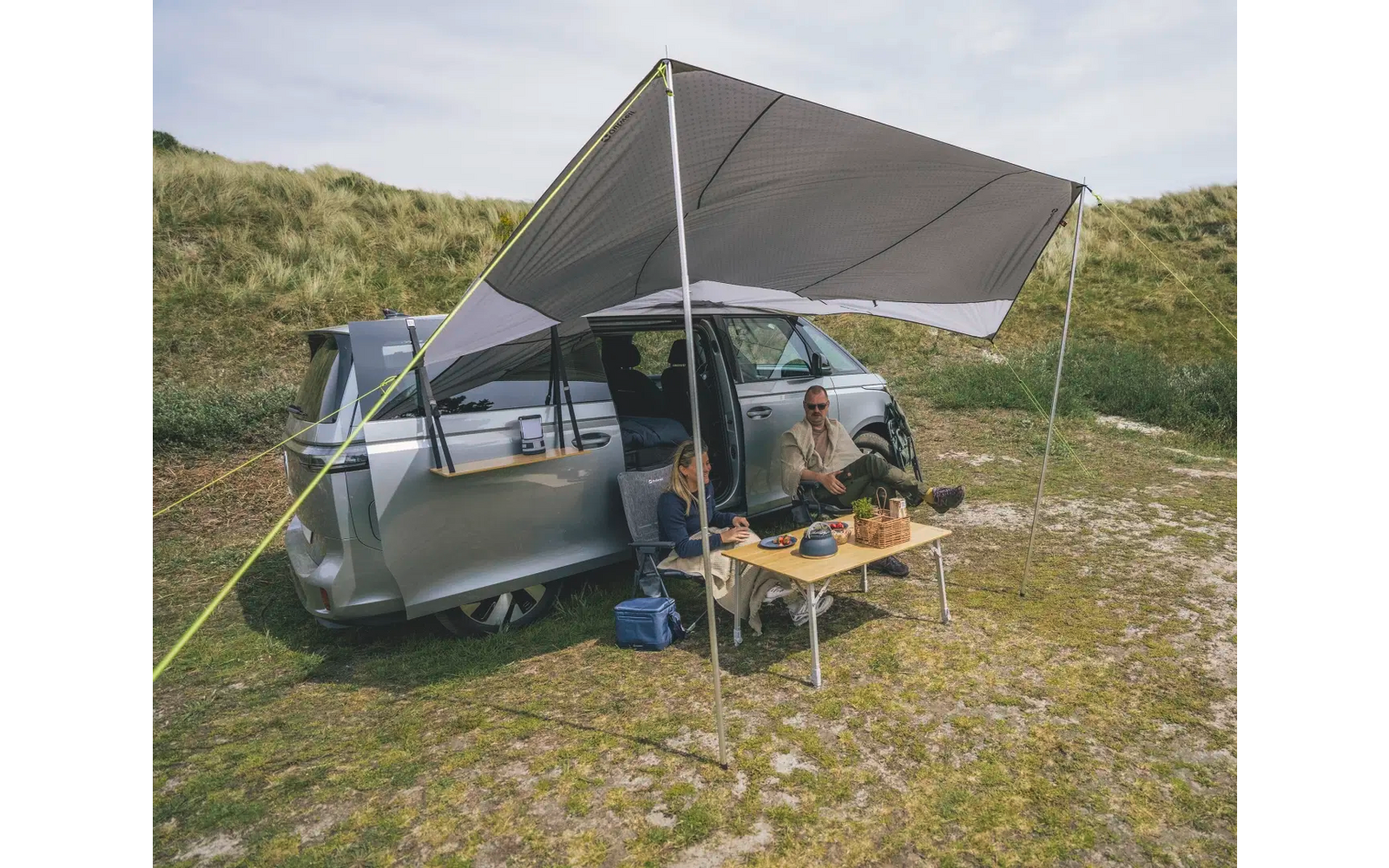 Wohnmobil steht auf einer Wiese mit geöffneter Markise und zwei Personen sitzen an einem Campingtisch