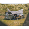 Campingfahrzeug mit zwei Personen, die unter einem Sonnensegel sitzen, vor grasbewachsenen Dünen