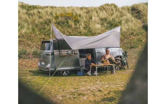 Campingfahrzeug mit zwei Personen, die unter einem Sonnensegel sitzen, vor grasbewachsenen Dünen