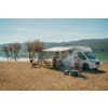 Wohnmobil mit ausgefahrener Markise an einem Seeufer; drei Personen bei Campingstühlen und Tisch, zwei Fahrräder, Grill mit Rauch, Bäume und Berge im Hintergrund