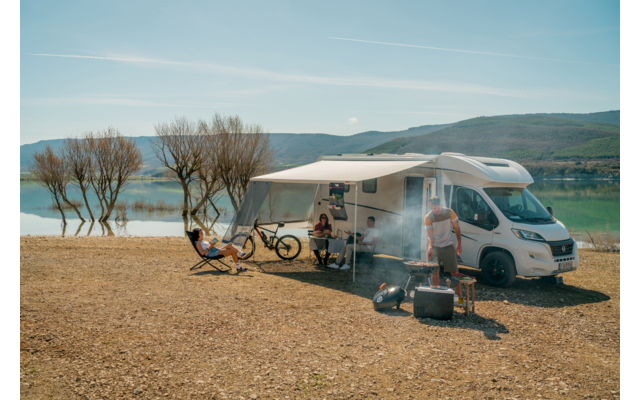 Wohnmobil mit ausgefahrener Markise an einem Seeufer; drei Personen bei Campingstühlen und Tisch, zwei Fahrräder, Grill mit Rauch, Bäume und Berge im Hintergrund
