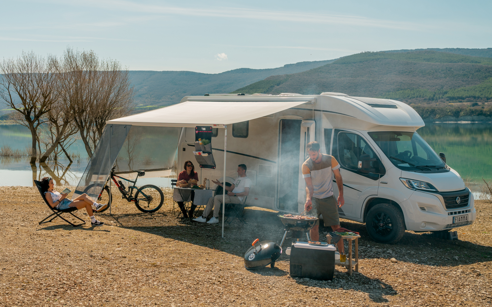 Wohnmobil mit ausgefahrener Markise an einem Seeufer; drei Personen bei Campingstühlen und Tisch, zwei Fahrräder, Grill mit Rauch, Bäume und Berge im Hintergrund