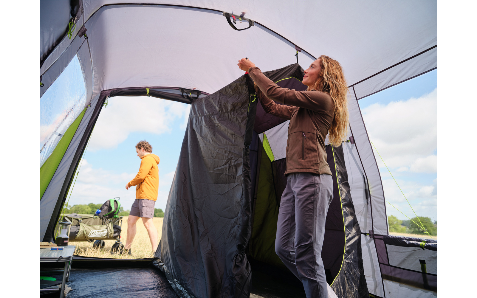 Zeltinnenraum, eine Person hält einen schwarzen Innenvorhang, eine weitere Person steht draußen in gelber Jacke, Campingstühle, grünes Feld und blauer Himmel mit Wolken