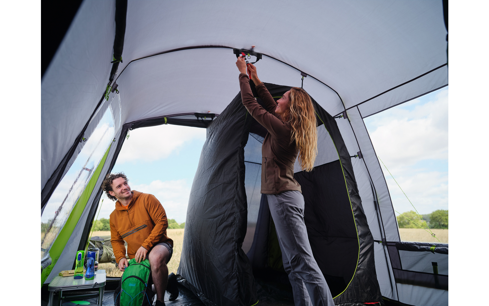 Zwei Personen in einem großen Campingzelt: eine Frau steht und hält ein schwarzes Stoffteil am Zeltfirst, ein Mann sitzt links auf einem Stuhl neben einem grünen Rucksack und einer Flasche; durch die Zeltöffnungen sind Wiese und Himmel sichtbar